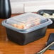 A black Genpak plastic container with a clear lid on a table with a black napkin and fork.
