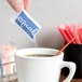 A hand holding an Equal sugar substitute packet over a white cup of coffee with a red straw.