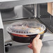 A hand opening a microwave with a black Choice plastic container with food inside.