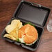 A sandwich with cheese and lettuce and a bag of chips in a black Genpak foam container.