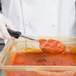 A gloved hand uses a Vollrath black oval portion spoon to scoop red sauce into a container.