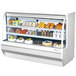 A Turbo Air white curved glass refrigerated deli case with food on shelves.