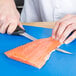 A person's hand using a Mercer Culinary Millennia Narrow Fillet Knife to cut salmon on a blue cutting board.