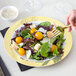 A hand holding a Visions white plastic plate with a gold lattice design over a salad with yellow tomatoes and cheese.