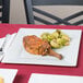 A Visions white plastic square plate with meat and brussels sprouts on a table.