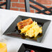 A Visions black plastic square plate with sausage and scrambled eggs on a table.