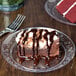 A Choice Crystal clear plastic plate with a slice of chocolate cake with chocolate sauce and whipped cream and a fork.