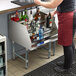 A woman in a red apron uses a Regency stainless steel liquor display rack behind a bar counter.