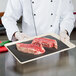 A chef holding a tray of raw meat on black steak paper.