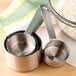 A Tablecraft stainless steel measuring cup and spoon set on a table with a bowl of flour.