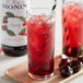 A glass of red cherry drink with ice and a straw with a bottle of Monin Premium Cherry Fruit Syrup.