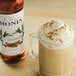 A Monin Premium Cinnamon Flavoring Syrup bottle next to a glass of coffee with whipped cream and cinnamon.
