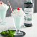 A glass of mint julep with a straw and whipped cream and a bottle of Monin Premium Green Mint Flavoring.