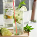 A glass of Monin mojito mix with sliced limes and mint leaves on a wooden tray.