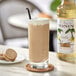 A glass of milkshake made with Monin Premium Vanilla Flavoring Syrup on a coaster with a cookie and a bottle of syrup.