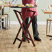 A person holding a Lancaster Table & Seating folding wood tray stand next to a table.