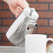 A hand pouring coffee from a silver stainless steel coffee pot into a white mug.