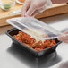 A person in plastic gloves holding a rectangular black plastic container filled with food with a black plastic lid.