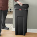 A man in a chef's uniform standing next to a Rubbermaid black square commercial trash can.