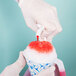 A person in white gloves pouring red Carnival King Watermelon snow cone syrup into a cup of snow.