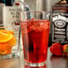 A Libbey pint glass filled with red liquid and ice on a table with a strawberry and a bottle of Jack Daniels.