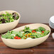 A Manila oval melamine bowl filled with salad on a table next to a stack of plates.