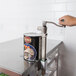 A hand using a mounted can opener to open a can of food on a stainless steel surface.