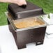 A person's hand opening a container of food in front of a Sterno Copper Vein Fold Away Chafer on a table in an outdoor catering setup.