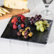 A black Madison Avenue faux slate display board with cheese and fruit on a table.