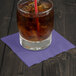 A glass of ice tea with ice and a straw on a purple Creative Converting beverage napkin.
