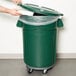 A person putting white trash in a green Rubbermaid BRUTE trash can with a green lid.