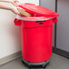 A woman opening a Rubbermaid red trash can with a lid to put a cardboard box inside.