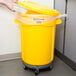 A person putting a white container into a yellow Rubbermaid commercial trash can.