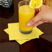 A hand holding a glass of orange juice on a yellow Creative Converting Mimosa Yellow beverage napkin.