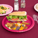 A Creative Converting hot magenta pink paper plate with a sandwich, chips, and other food on a table.