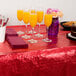 A table with a red Creative Converting plastic table cover and glasses of orange juice and food.
