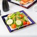 A Fineline white plastic plate with blue rim and gold bands holding salad with tomatoes and cucumbers.