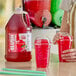 A gallon jug of Carnival King cherry slush syrup concentrate next to two cups of red cherry slushies with dome lids.