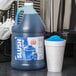 A blue Carnival King jug of slushy syrup next to a cup of blue slushy with a blue lid.