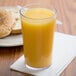 A Cambro clear plastic tumbler filled with orange juice next to a bagel on a table.