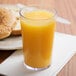 A clear plastic Cambro tumbler filled with orange juice next to a bagel on a table.