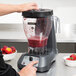 A person using a Hamilton Beach food blender to blend a bowl of strawberries, peaches, and other fruit into a red smoothie.
