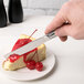 A hand holding a Libbey stainless steel utility/dessert knife cutting a piece of cheesecake with cherry topping.