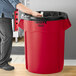 A man standing next to a red Rubbermaid BRUTE trash can.