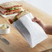 A hand holding a sandwich in a white paper bag.
