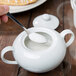 A person using a spoon to add sugar to a white 10 Strawberry Street porcelain sugar bowl.