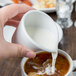 A person pouring milk from a Libbey Reflections porcelain creamer into a white cup of coffee.