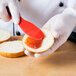 A person in gloves is spreading jam with a red HS Inc. polypropylene sandwich spreader on a piece of bread.