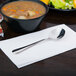 A Libbey stainless steel bouillon spoon on a white napkin over a bowl of soup.