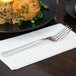 A stainless steel Libbey Aegean dessert fork on a white napkin next to a plate of food.